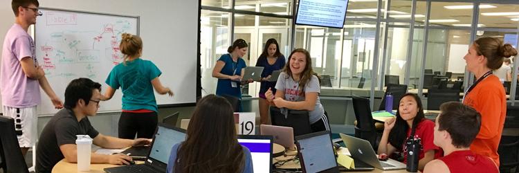 A lively classroom scene: several students sit and stand around a large round table, while one student writes on a white board and two others stand together holding laptops the background.