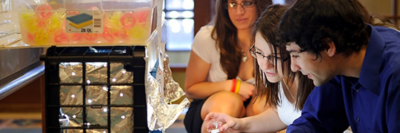 Cropped photo of three students gathered around a device in a research lab. 