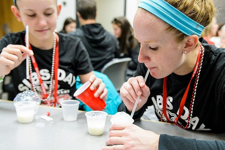 Two students use straws to blow bubbles into small cups as part of a science activity.