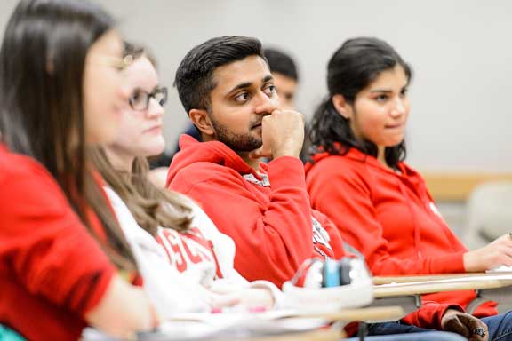 Several students sit at desks in a classroom, listening to a lecture.