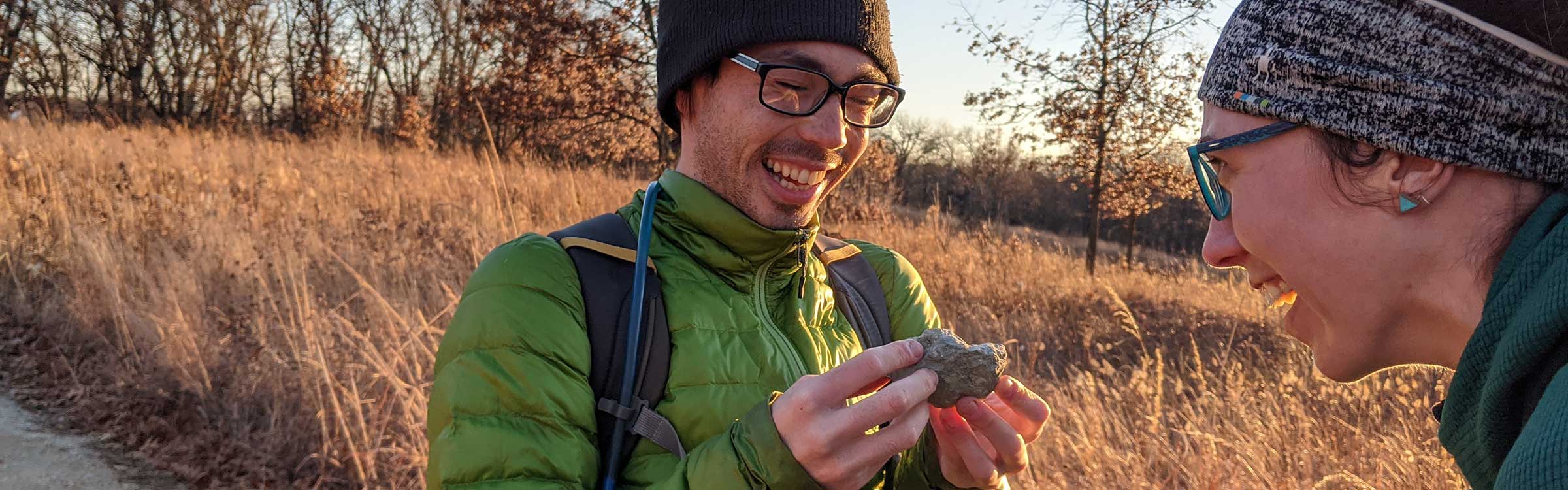 Two students are in a prairie wearing warm hats and jackets. One holds up a rock to show the other. 