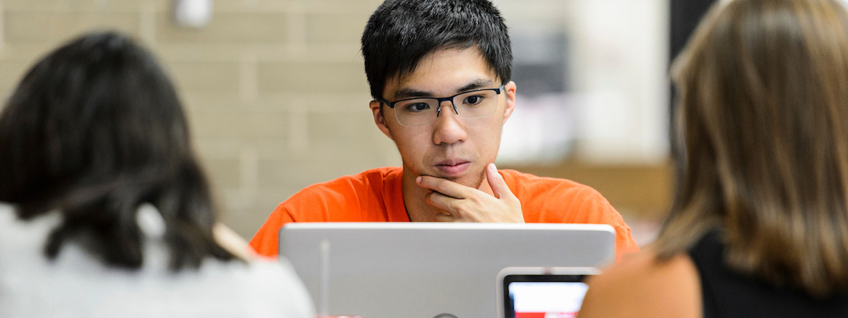 A student sitting at a table looks at his laptop with his chin in his hand. Two students sit at the other side of the table, their backs to the camera.