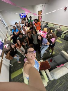 One student holds a camera for a selfie with a large group of students gathered in a room with pink streamers and balloons.