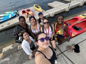 Seven students stand and smile next to a dock filled with kayaks next to a lake.