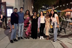 Seven summer research students pose together on a sidewalk outside of a restaurant at night.