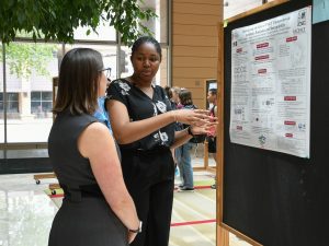 Danielle Amaegbo, a summer research student, stands in front of her research poster, gesturing with her hands as she explains her research to a listening woman.