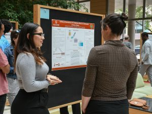 A student with long, dark-brown hair and glasses stands next to a research poster, discussing her research with a visitor. 