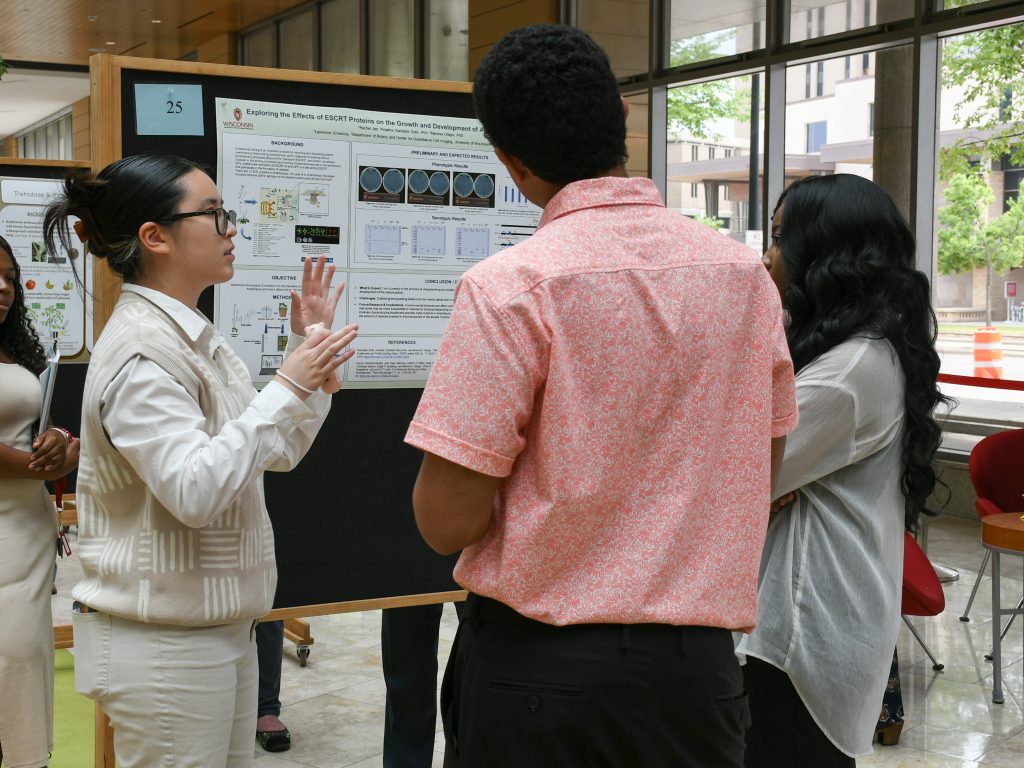 One student gestures with her hands while explaining her research poster to two attentive students.