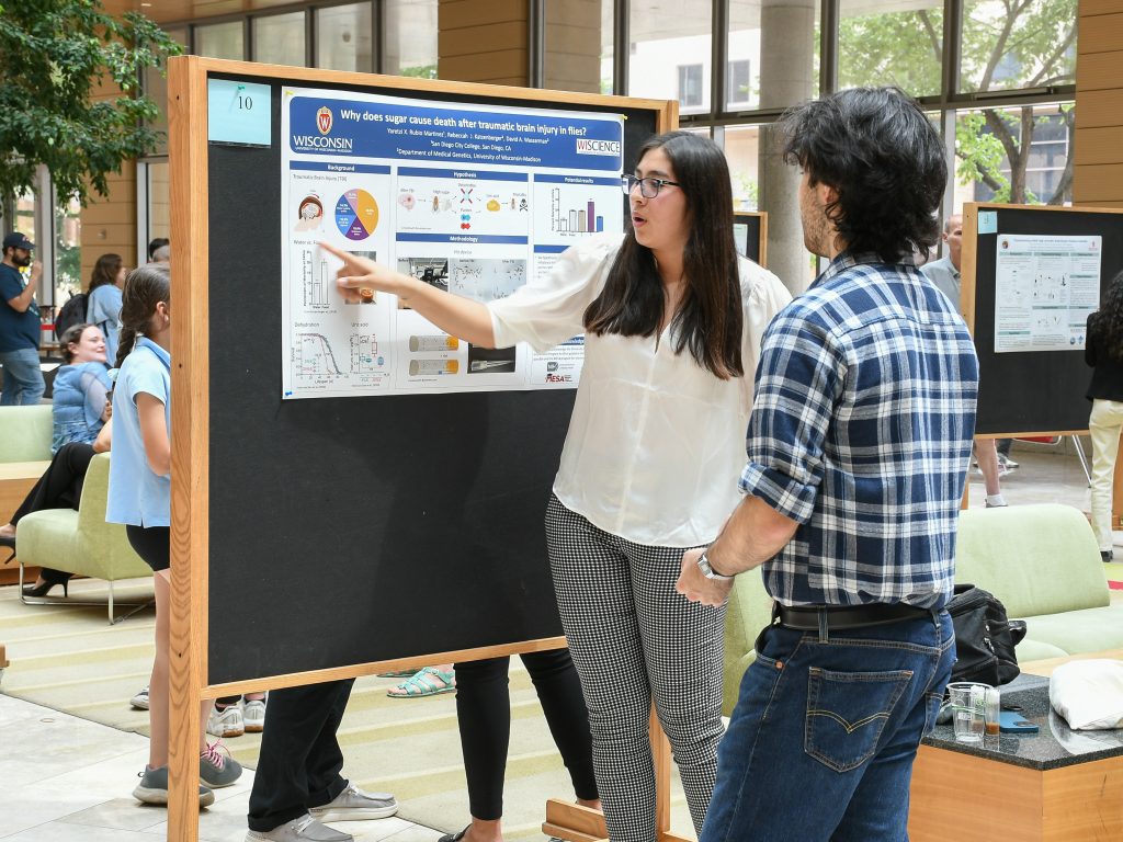 A student points to her research poster, explaining her research to another student. 