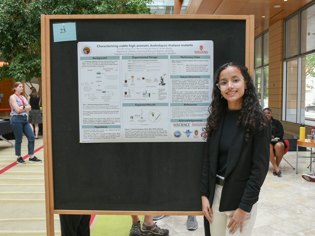 A smiling student with long, dark-brown hair and glasses stands next to a research poster. 