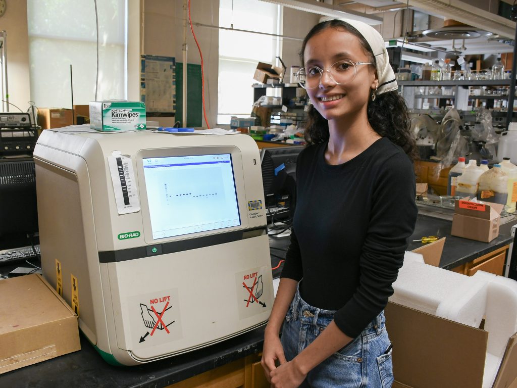 A student wearing glasses, an off-white hair scarf, and black shirt stands smiling next to a machine in a lab. 
