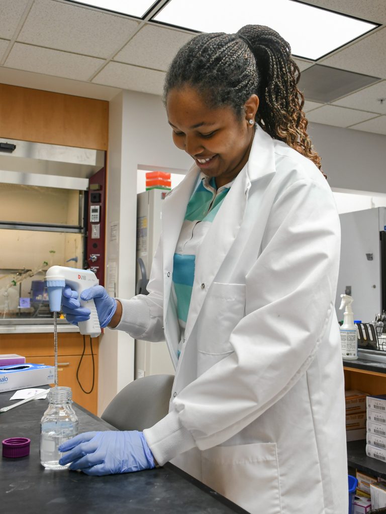 Danielle Amaegbo, a summer research student, stands at a lab bench wearing a lab coat and holding a pipette controller.