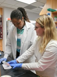An undergrad student and her grad student mentor are wearing white lab coats and looking at something on a lab bench. 