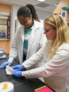An undergrad student and her grad student mentor are wearing white lab coats and looking at something on a lab bench.