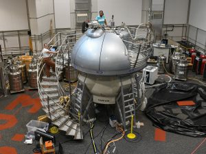 Inside the National Magnetic Resonance Facility: a large room with a tall machine that has a curved staircase around the side, leading to a domed top. One student stands at the top of the machine while another stands on the stairs.