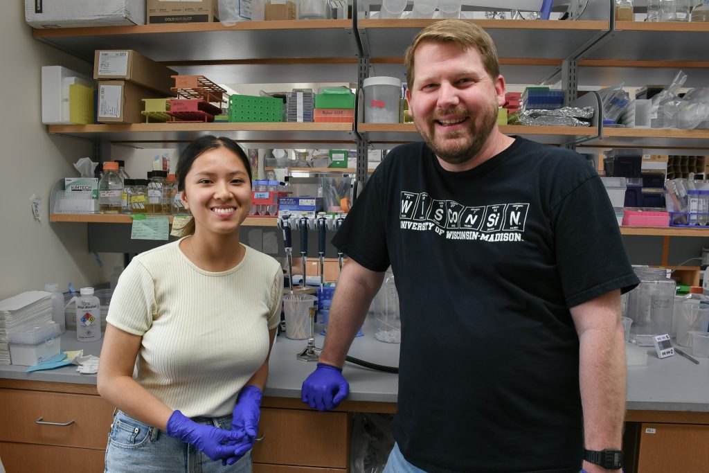 A student and a professor stand and smile in front of a lab bench.