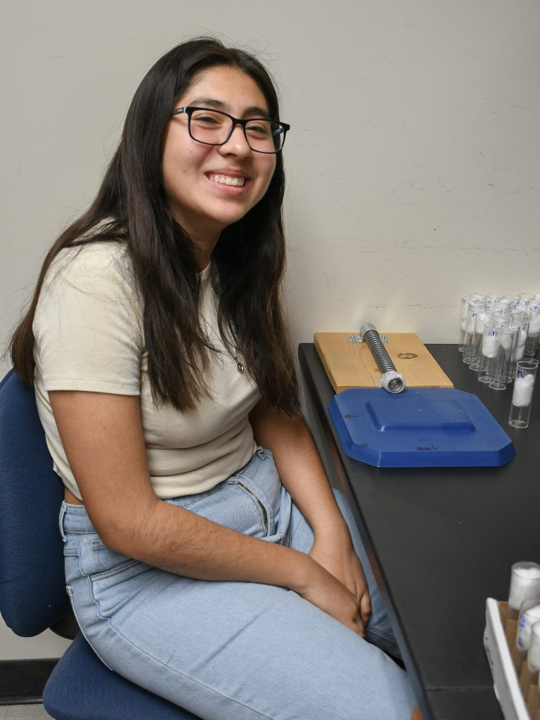 A student sits at a lab bench, smiling. On the bench in front of her is a spring on a wooden board: the High-Impact Trauma (HIT) device she's using as part of her research project.