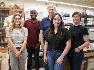 Six members of the Hite Lab stand together and smile with lab equipment in the background. 