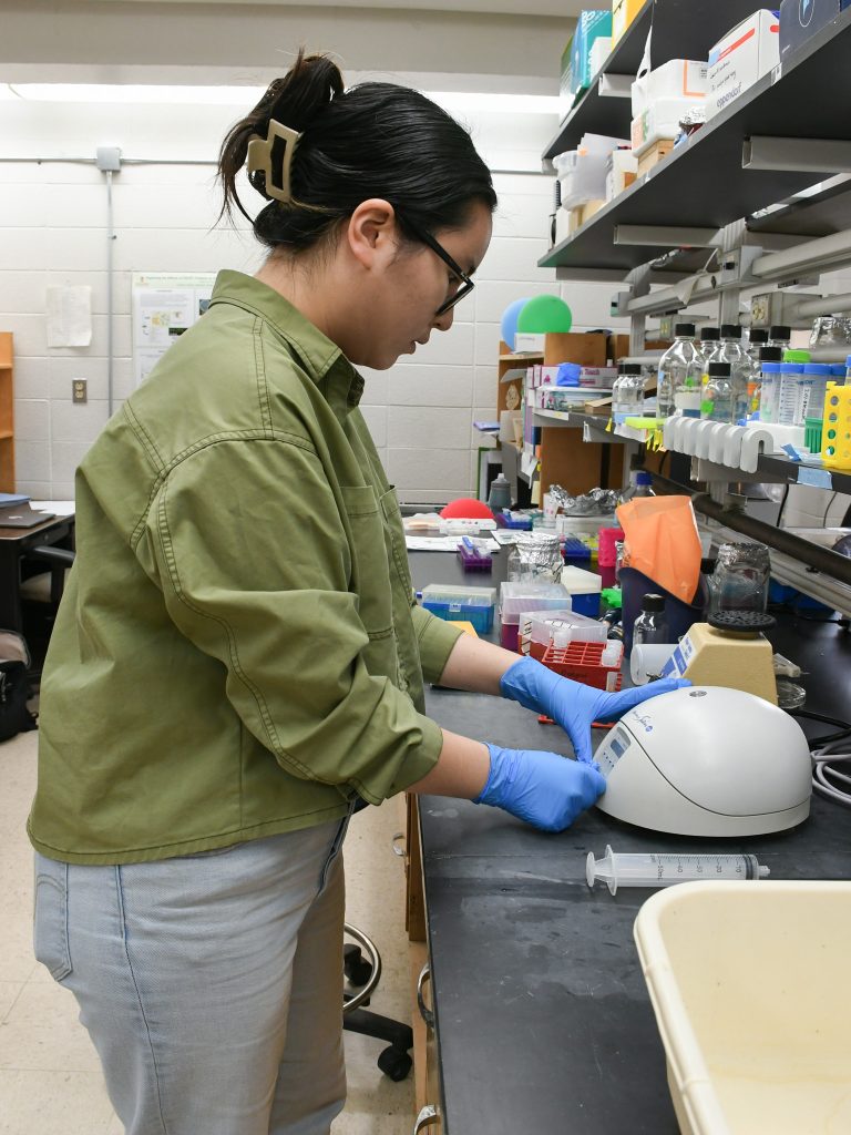 A student wearing a green jacket and lab gloves places a sample in a machine on a lab bench.