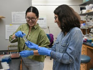 Rachel Joo, a summer research student, hands a test tube to her mentor, Ariadna Gonzalez Solis, who is grabbing onto it and smiling. They are in a lab. 