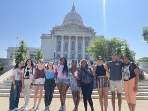 Ten students with linked arms stand and smile in front of the Wisconsin State Capitol, a large white building with columns and a dome. 