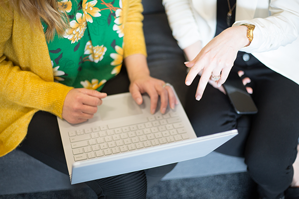 Two women are seated on a couch; one holds a laptop while the other points to something on the computer screen.