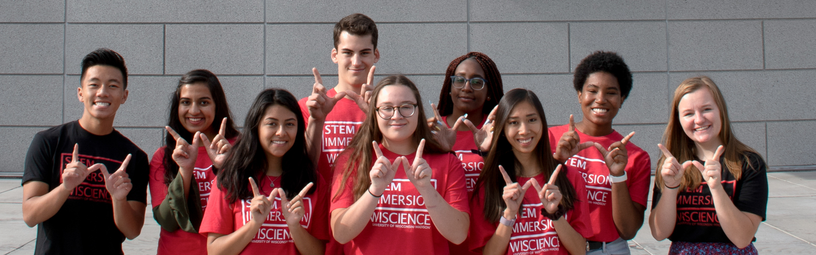 A group of nine students smile and make the "W" sign with their hands. They are wearing T-shirts that read "STEM IMMERSION - WISCIENCE."