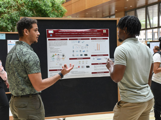 A man with short, curly brown hair gestures at a poster on a bulletin board while another man with ear-length black hair listens.