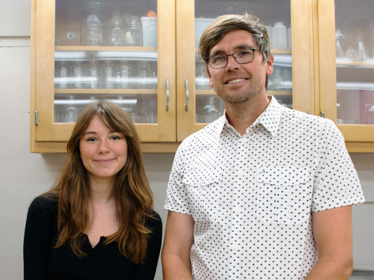 A woman with long brown hair and a man with short hair and glasses stand in front of a cabinet of lab equipment. 