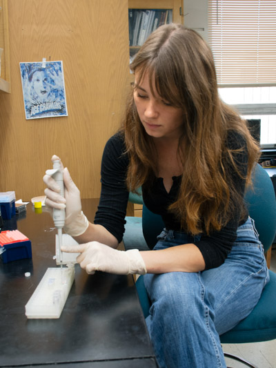A young woman with long brown hair sits at a lab bench, using a pipette. 
