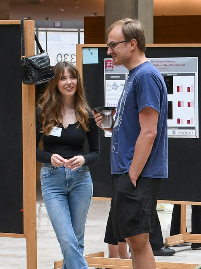 A woman with long hair smiles while a man with short hair and glasses looks at a bulletin board in front of them. 