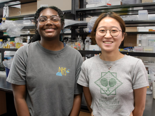 Two smiling women stand together in front of a lab bench, one with dark, curly hair, glasses, and a dark gray t-shirt, and the other with straight brown hair, glasses, and a light gray t-shirt. 