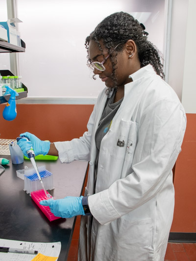 A young woman with dark, curly hair wears a white lab coat and uses a pipette at a lab bench. 