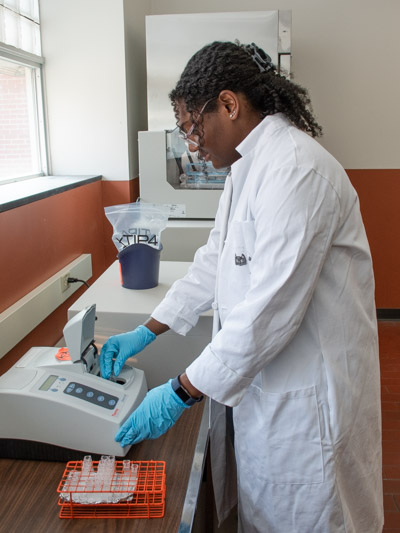 A young woman with dark, curly hair wearing a white lab coat places something in a device in a science lab. 