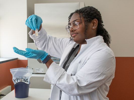 A young woman with dark, curly hair wears a white lab coat and blue latex gloves, holding a small vial of liquid. 