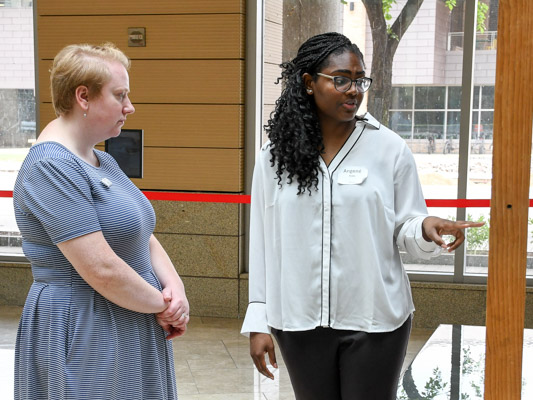 A young woman with long, dark hair and a white shirt talks about her research to a woman with short, light hair. 