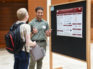 Standing in front of his poster at the CMBS Poster Fair, student Adam Ghazzali smiles while explaining his research to another student.
