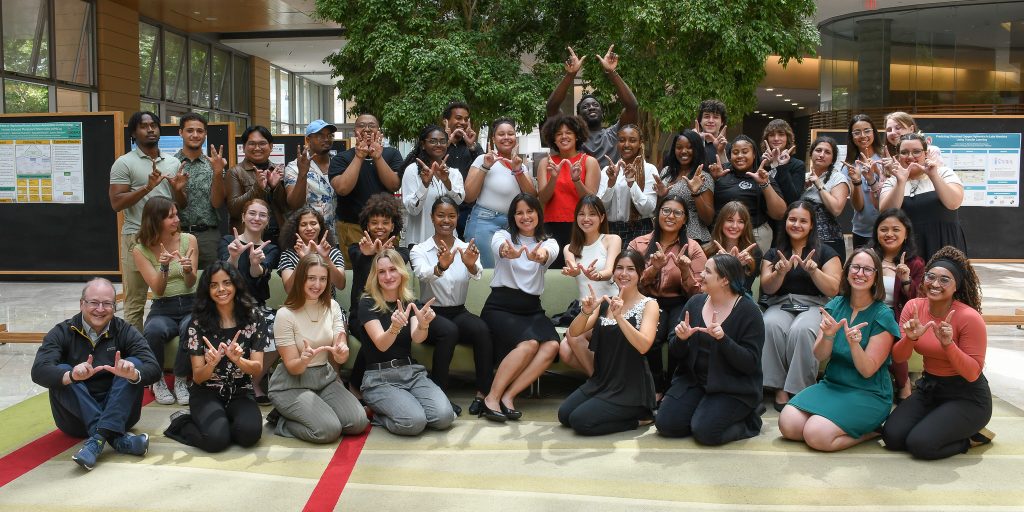 About 30 students from the 2024 CMBS Summer Research Program plus the program directors pose together in an atrium, making the "W" sign with their hands.