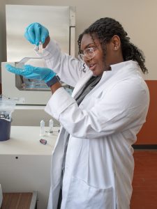 CMBS student Angene' Hale wears a lab coat and gloves and holds a vial of liquid as she works on her research project in the Huynh Lab.