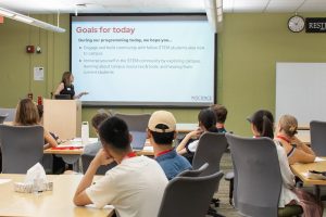 Sarah Sweger, Assistant Director for STEM Student Engagement, stands at a podium and reads from a projector screen. In front of her, several students sit at tables and listen.