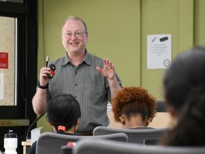 Professor David Wassarman gestures with his hands as he gives a presentation about the research process to the 2023 Summer Research Program cohort. Several students are seated in front of him.