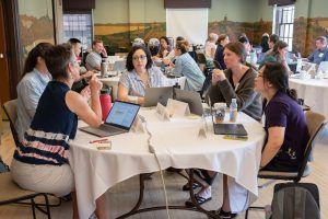 Five biology educators sit around a table with laptops, and behind them are several other tables with other educators. 
