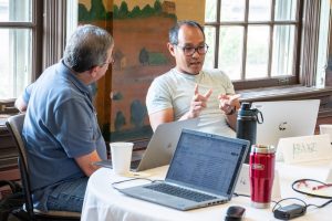 Sitting at a table with laptops, one biology educator listens as another talks. 