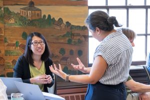 One biology educator, stands and gestures with her hands as she talks to another educator, who is seated and smiling. 