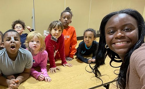 A student from the class Service with Youth in STEM smiles at the camera in front of a table of elementary school students. 