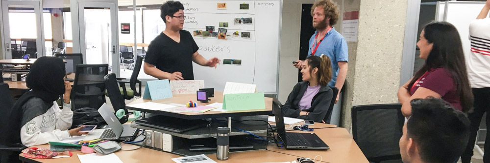 Five students stand and sit around a table and whiteboard in a classroom.