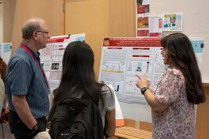 Summer research program student Elaine Torres-Lopez stands next to her presentation poster, talking and gesturing with her hands as two other people listen.