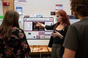 Eleanor Ford, a summer research program student, stands in front of her research poster, pointing at it while talking to two people. 