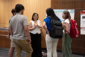 Alexia S. Meléndez Rivera, a summer research student, stands next to her research poster with a group of four people, listening as a visitor asks her a question. 