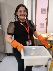 Alexia S. Meléndez Rivera, a summer research student, smiles while wearing orange heat-resistant gloves and holding a tray that contains a beaker of hot liquid. 